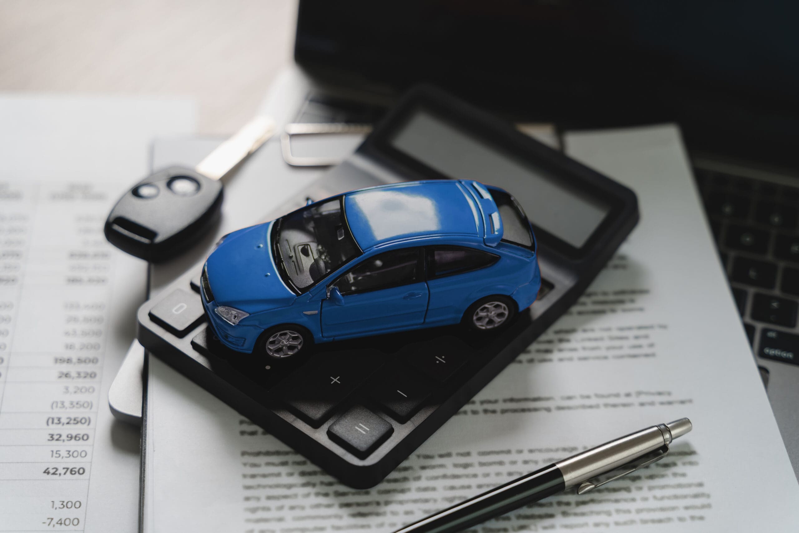 Blue toy car model on calculator with financial document showing numbers and key, pen and laptop on desk, representing car finance price calculation, auto loan value assessment, vehicle cost estimation, budget planning concept