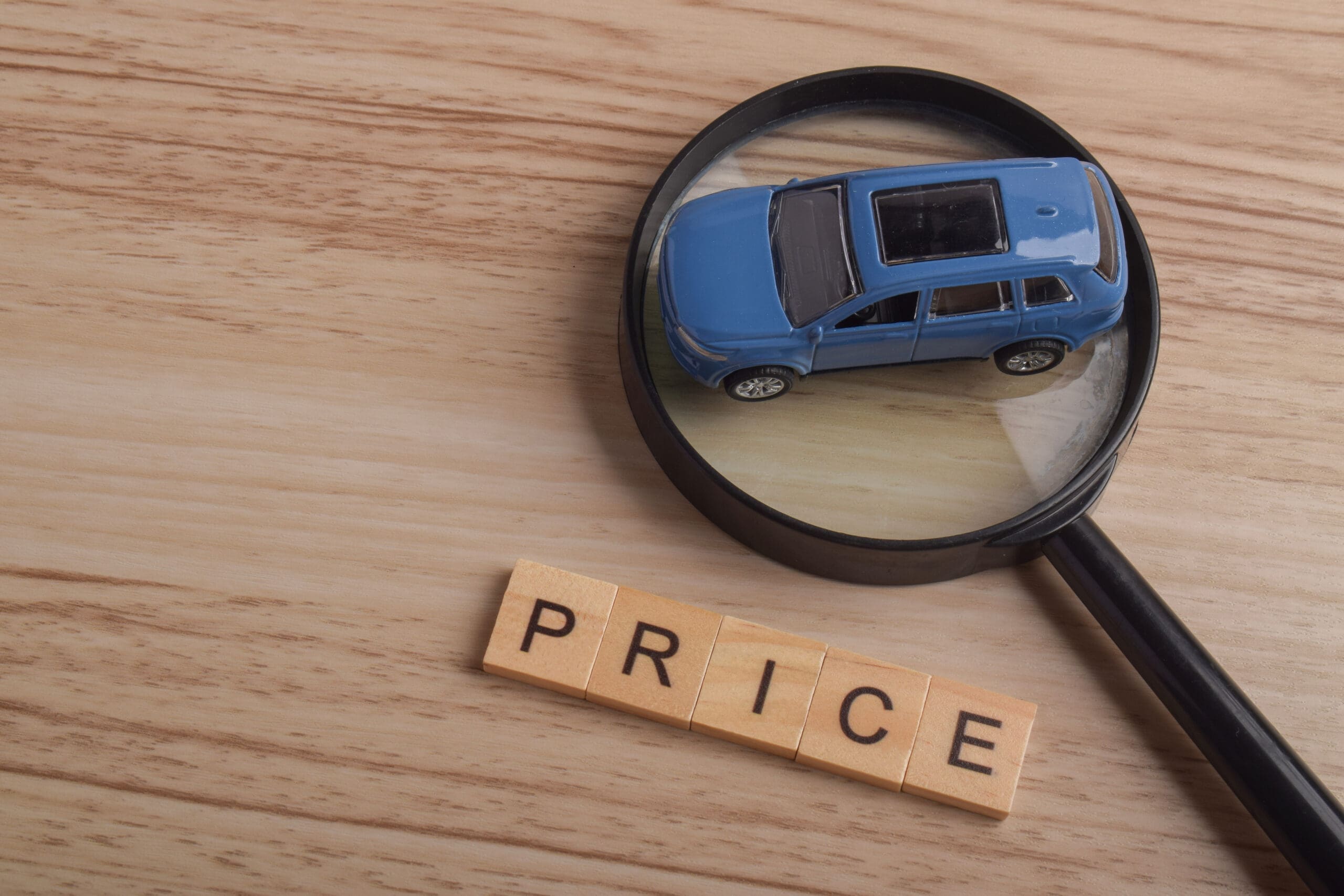 Magnifying glass highlighting toy car beside price lettering on wooden background, representing vehicle valuation, price comparison, automotive market research, car buying decisions, and cost analysis
